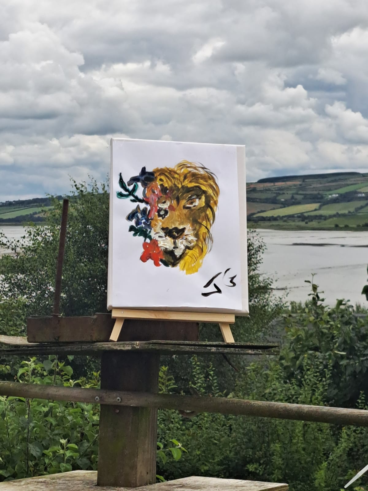 Image of a canvas with a painting of a lion against a background showing the Irish countryside, with a hill and lake and cloudy sky 